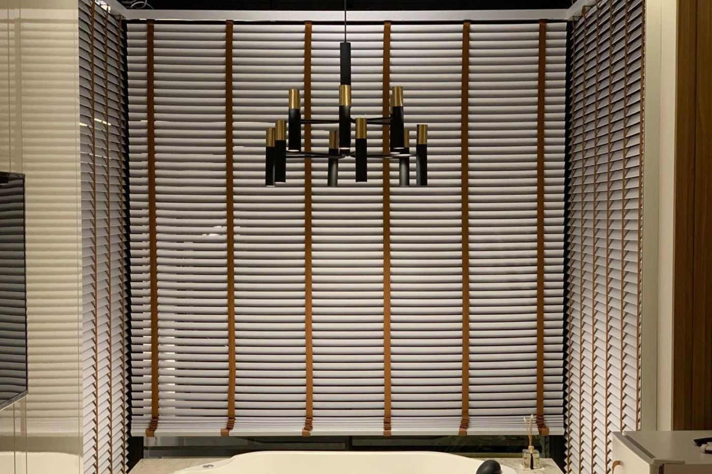 Persiana horizontal de madeira com tiras de couro caramelo em janela ampla de banheiro sofisticado, lustre pendente preto e dourado com múltiplos braços ao centro, luz suave difusa pelas lâminas, banheira e bancada de mármore claro visíveis abaixo, ambiente contemporâneo elegante, fotografia realista de interiores