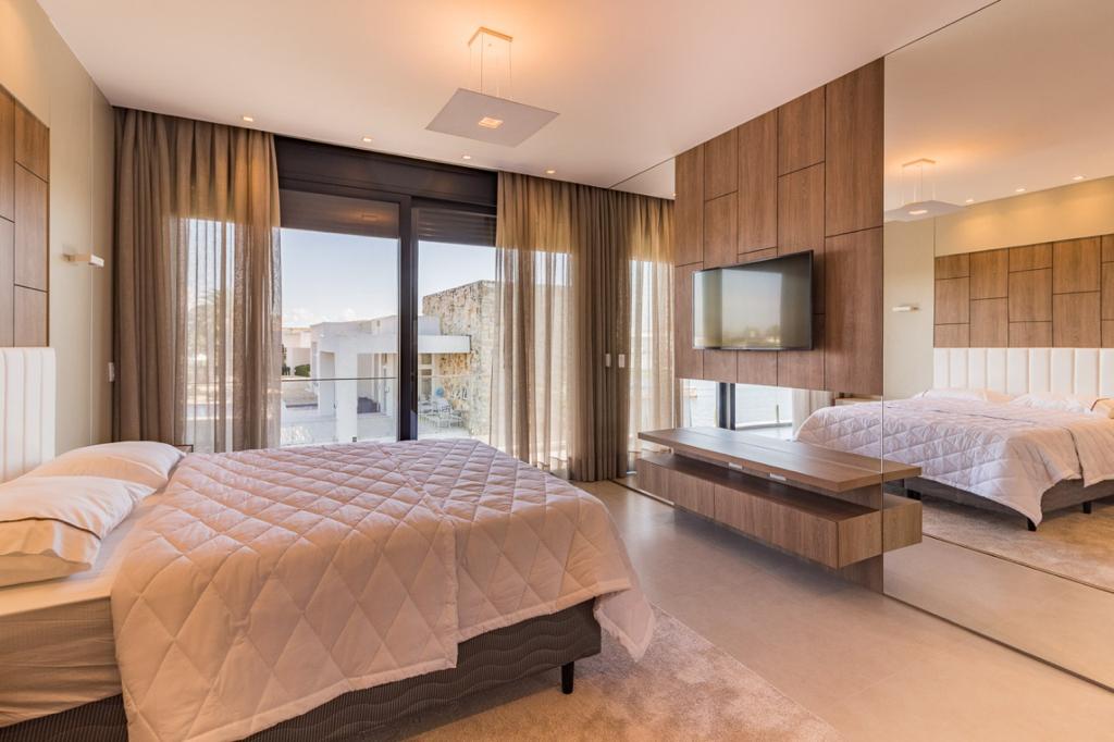 Quarto de casal amplo e elegante com cortinas de linho bege do piso ao teto emoldurando porta de vidro para varanda com vista para o mar, cama de casal com edredom acolchoado branco, painel de TV em madeira com espelho, luz dourada do fim de tarde, fotografia realista de interiores