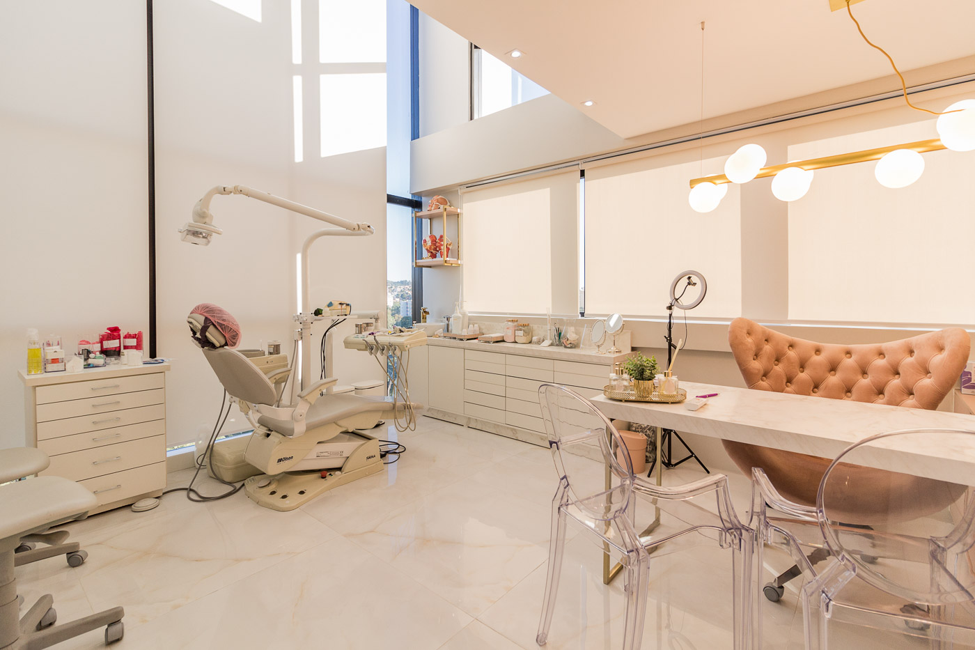 Consultorio clinico clean com cortinas roll-up bege translucidas em janelas amplas de pe direito duplo, poltrona odontologica, bancada branca com produtos, mesa de marmore com cadeiras acrilicas transparentes, iluminacao decorativa dourada, ambiente contemporaneo, fotografia realista de interiores