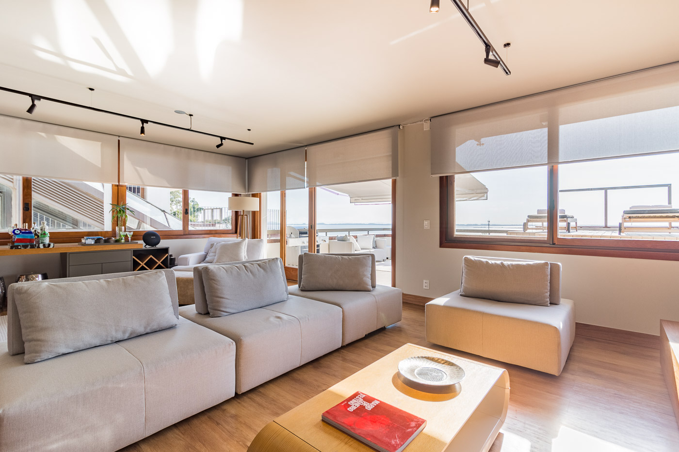 Sala de estar com cortinas roll-up translucidas em janelas amplas com vista para o mar e areas de lazer, sofas modulares cinza claro com almofadas, mesa de centro de madeira com revista, piso de madeira clara, iluminacao de trilho, ambiente contemporaneo, fotografia realista de interiores