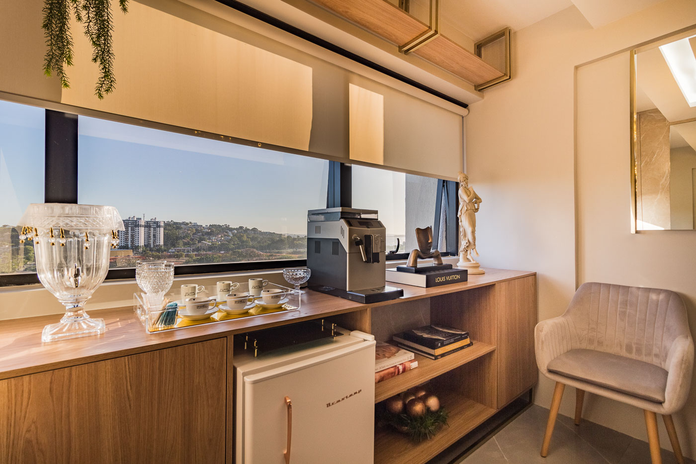 Bar planejado de madeira com cortina roll-up bege translucida em janela panoramica com vista para a cidade, cafeteira, tacas de cristal e frigobar embutido, poltrona estofada bege, decoracao sofisticada, luz natural quente, fotografia realista de interiores