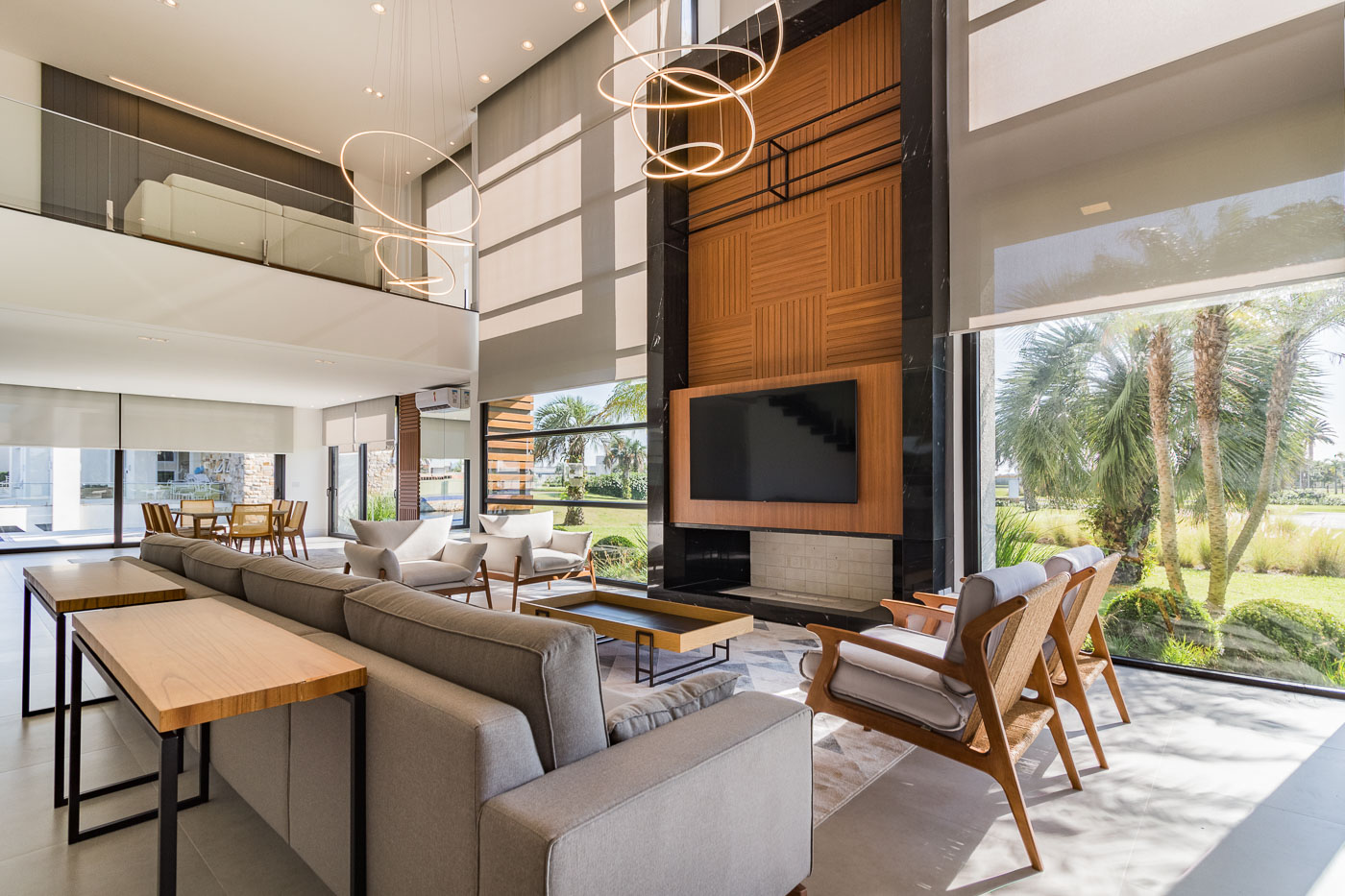 Sala de estar ampla com pe direito duplo, cortinas roll-up bege translucidas em janelas amplas do piso ao teto, painel de TV em madeira ripada e marmore preto, lustres circulares de LED suspensos, sofa cinza modular e poltronas de madeira, vista para jardim tropical, luz natural abundante, fotografia arquitetonica realista