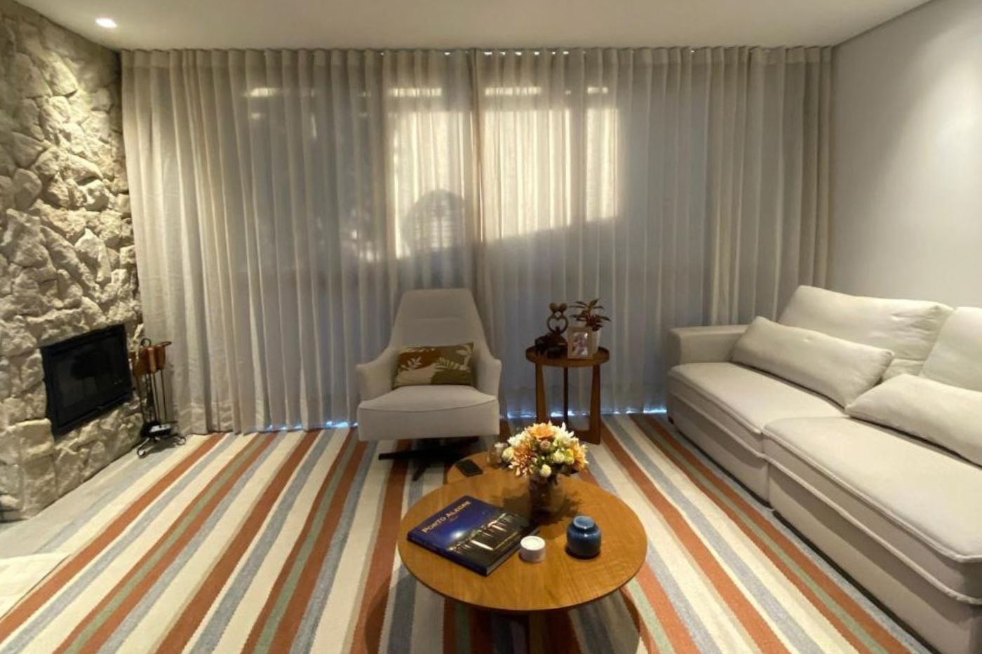 Sala de estar elegante com cortinas de tecido tradicional em tom claro do piso ao teto, sofás brancos e poltrona estofada, tapete listrado colorido, mesa de centro redonda de madeira com vaso de flores, luz natural suave, decoração contemporânea aconchegante, fotografia realista de interiores