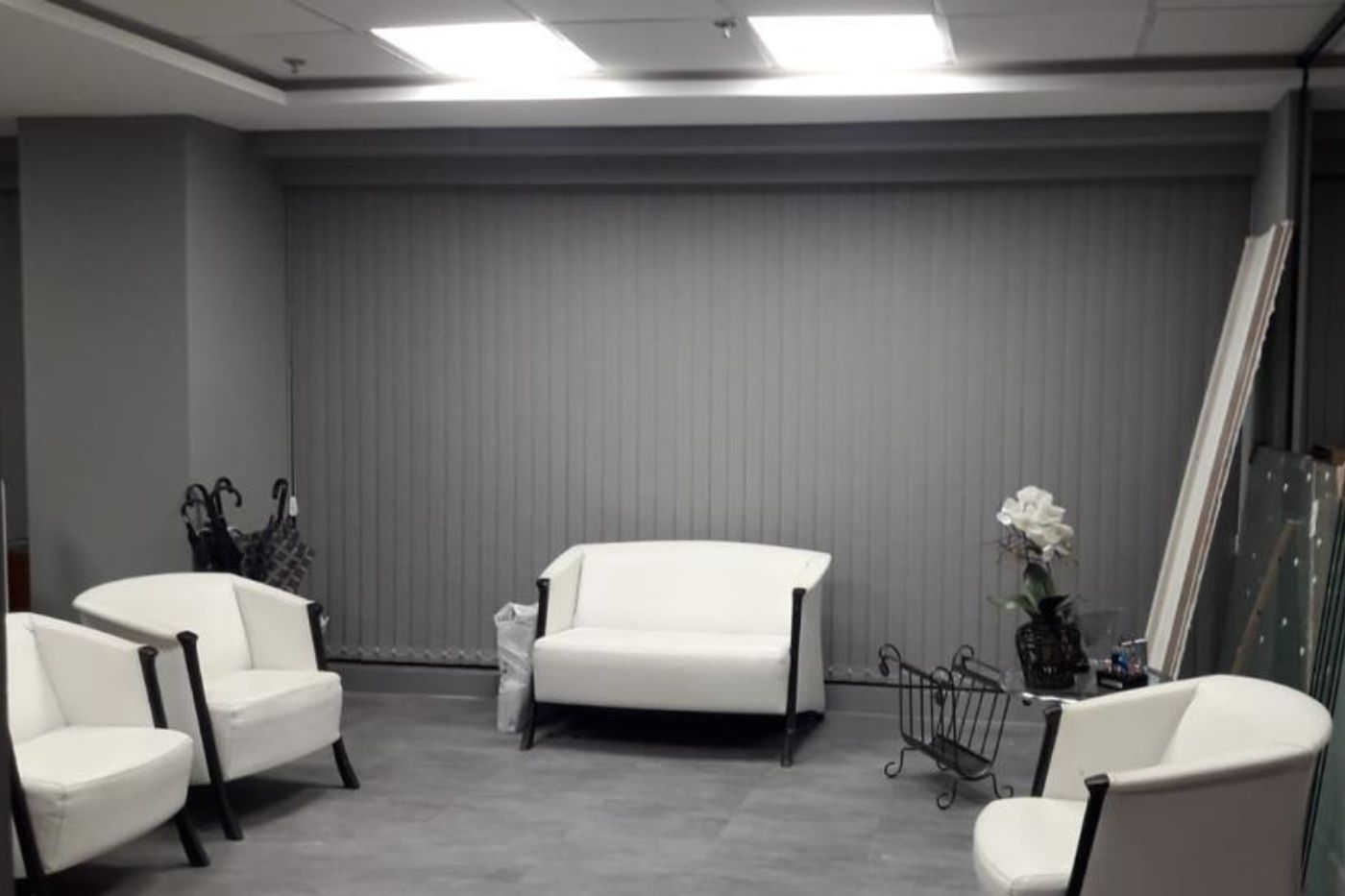 Sala de espera corporativa moderna com persiana vertical de lâminas cinza cobrindo ampla janela, poltronas e sofá em couro branco, piso em porcelanato acinzentado, luz natural difusa pelas lâminas, ambiente clean e sofisticado, fotografia realista de interiores