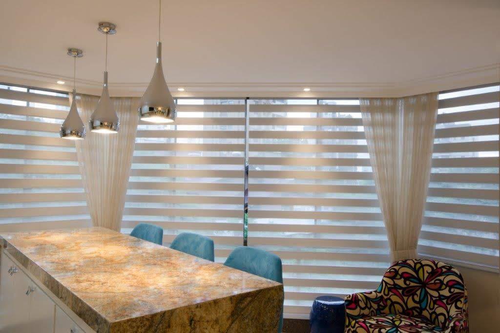 Sala de jantar moderna com cortinas double vision em tom bege claro nas janelas amplas, luz natural entrando em faixas horizontais suaves, mesa de granito com cadeiras estofadas azul-turquesa, poltrona estampada colorida, iluminação pendente moderna, fotografia realista de interiores