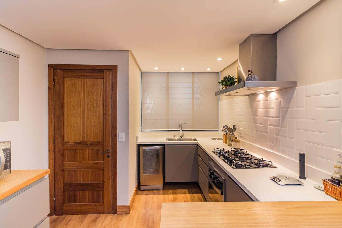 Cozinha planejada com cortina roll-up translucida em janela sobre a pia, coifa de inox suspensa, cooktop a gas, bancada branca, piso de madeira clara e porta de madeira macica, iluminacao embutida, fotografia realista de interiores