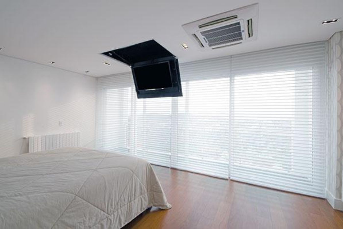 Quarto de casal com persiana horizontal branca cobrindo porta de vidro para varanda, ar-condicionado split de teto, cama de casal com edredom bege, piso de madeira, ambiente clean e luminoso, fotografia realista de interiores