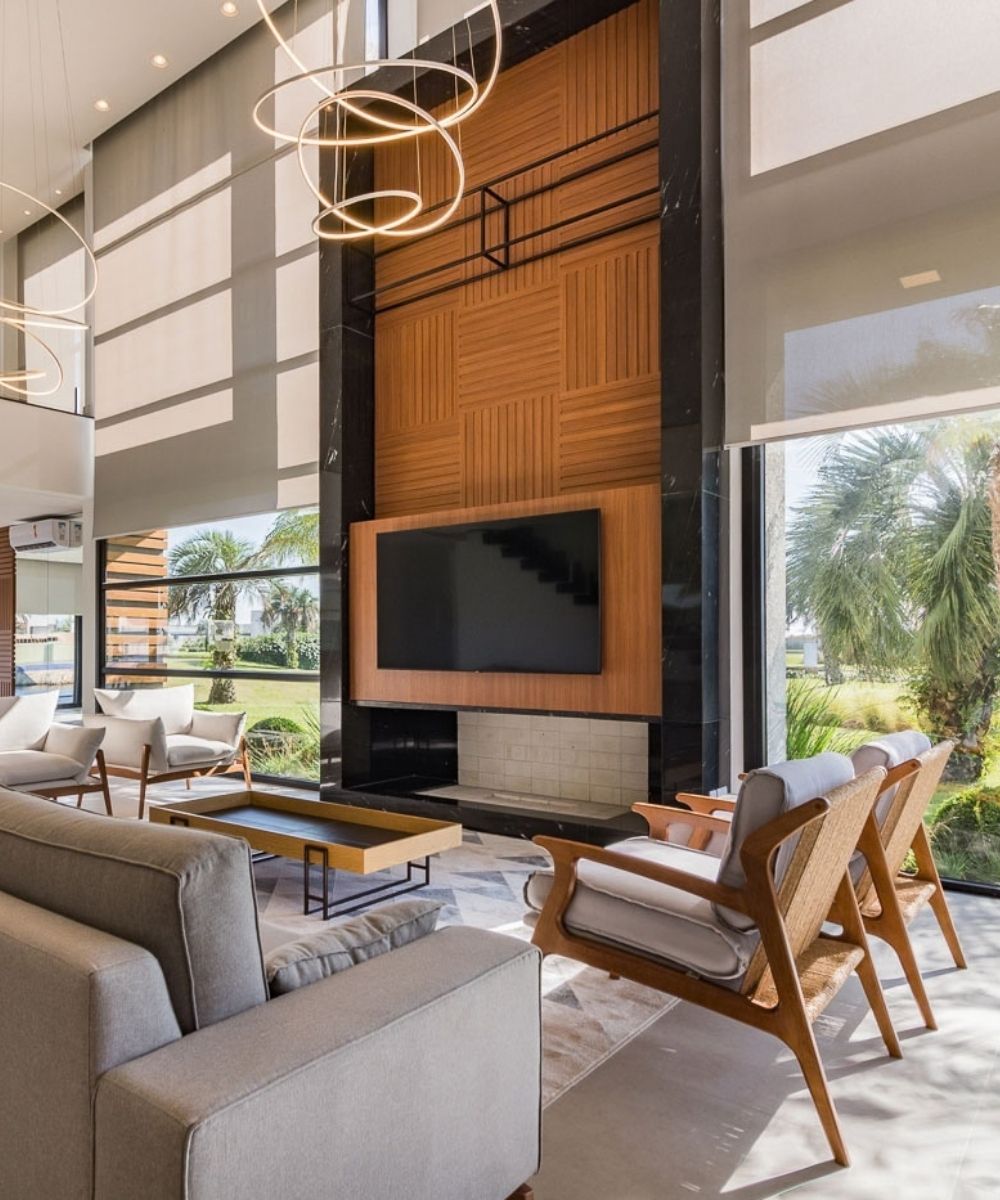 Sala de estar de pé-direito duplo sofisticada com painel de TV em madeira ripada e mármore preto, lustres circulares de LED suspensos, cortinas rolô bege translúcidas em janelas amplas do piso ao teto com vista para jardim tropical, sofás e poltronas cinza de design moderno, luz natural abundante, ambiente contemporâneo de alto padrão, fotografia realista de interiores