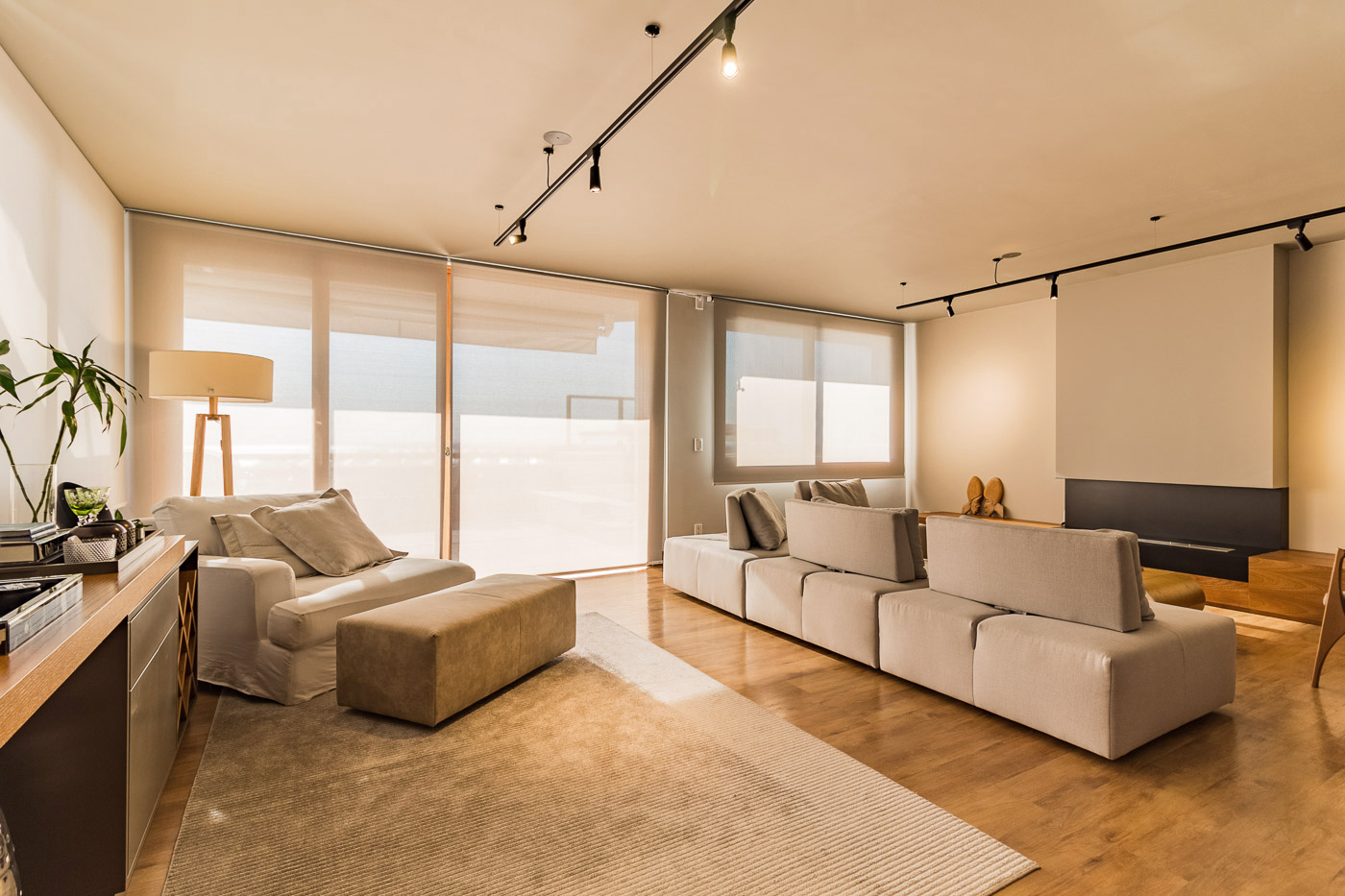 Sala de home theater com cortinas roll-up translucidas em portas de vidro para varanda, sofas modulares bege, tela de projecao retratil embutida no teto, piso de madeira clara, tapete bege, iluminacao de trilho, ambiente contemporaneo, fotografia realista de interiores