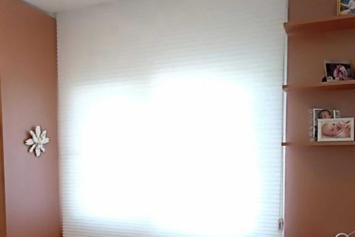 Persiana celular com estrutura em favo de mel tom branco em janela ampla de quarto com prateleiras cor terracota e enfeites decorativos, luz suave difusa criando efeito de brilho intenso na janela, ambiente aconchegante, fotografia realista de interiores